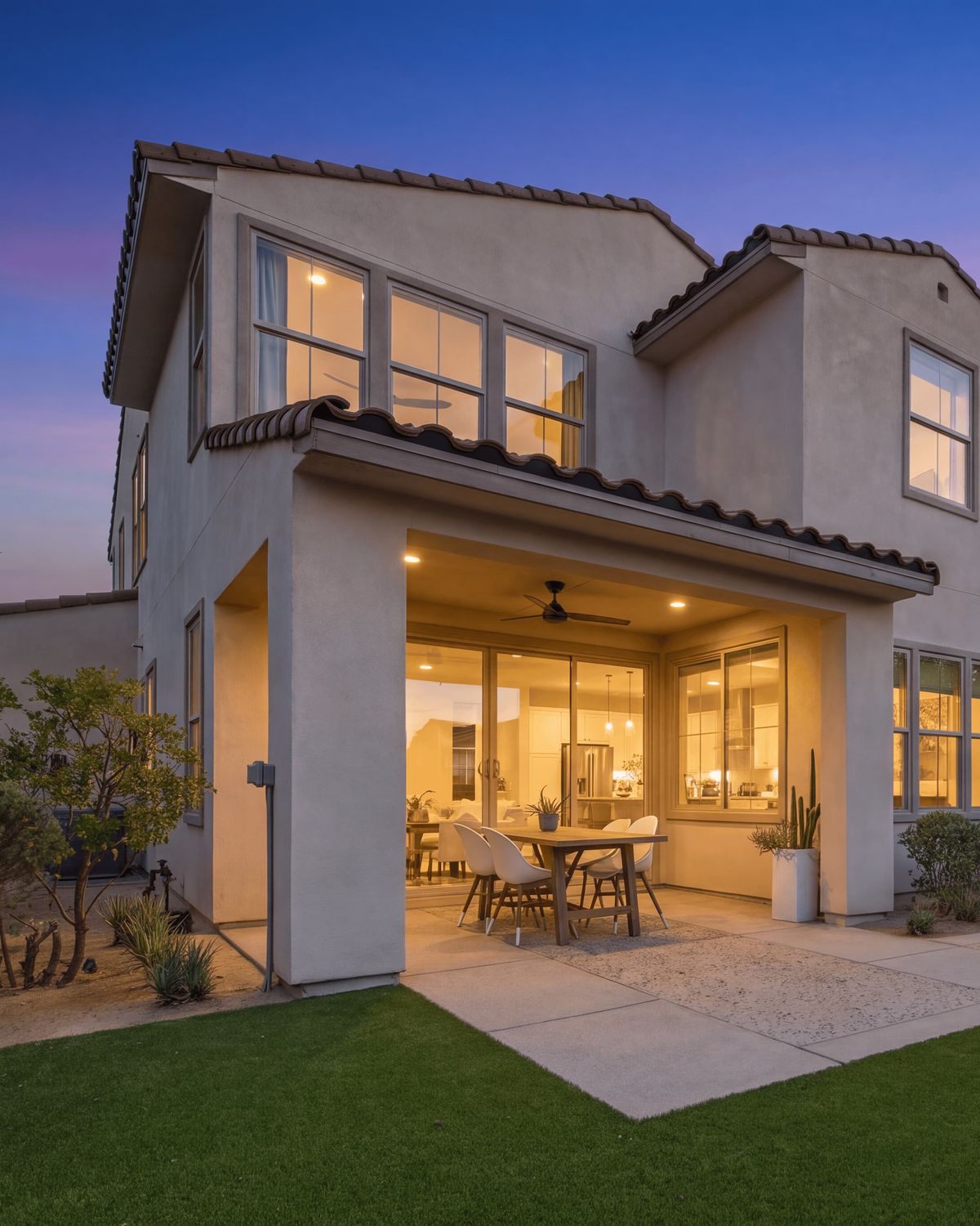 Two-story home backyard at dusk with warm glowing windows and a purple-pink sky